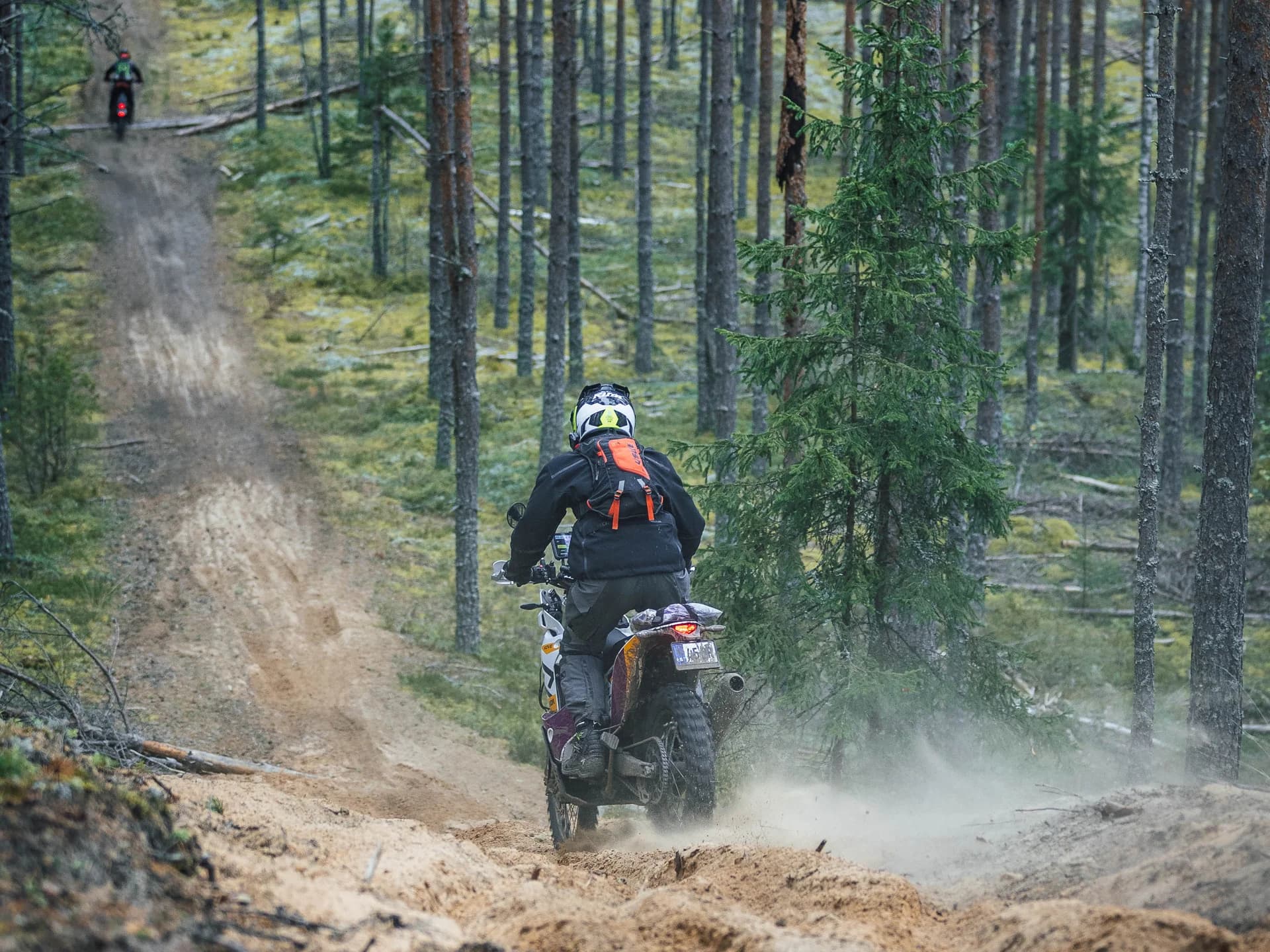 Enduro rider navigating a challenging sandy uphill trail through dense pine forest with dust clouds