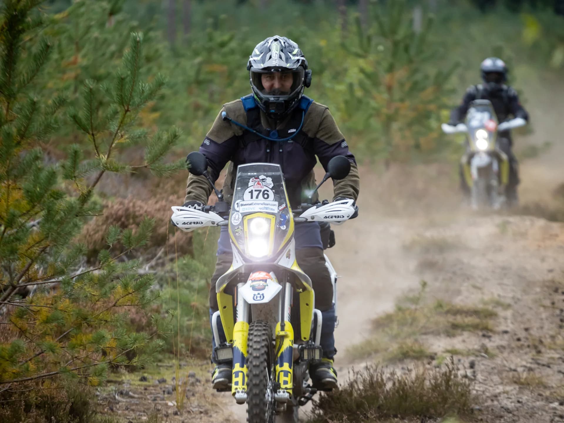 Two enduro riders on yellow and white motorcycles navigating a sandy trail through pine forest